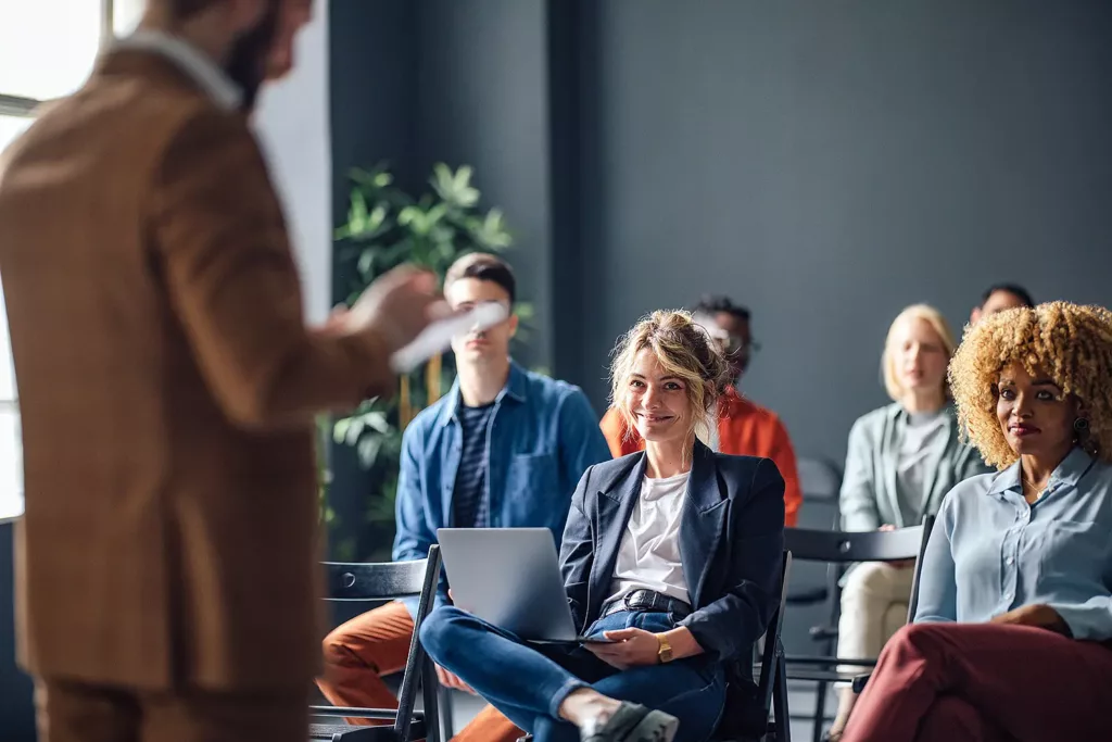 In a seminar room, a group of young adults of different origins can be seen sitting on black chairs. A man in the foreground on the left side of the picture seems to be ticking off a list. The focus is on a young woman who is wearing a dark blazer and loosely balancing a laptop on her legs. She smiles at the man.