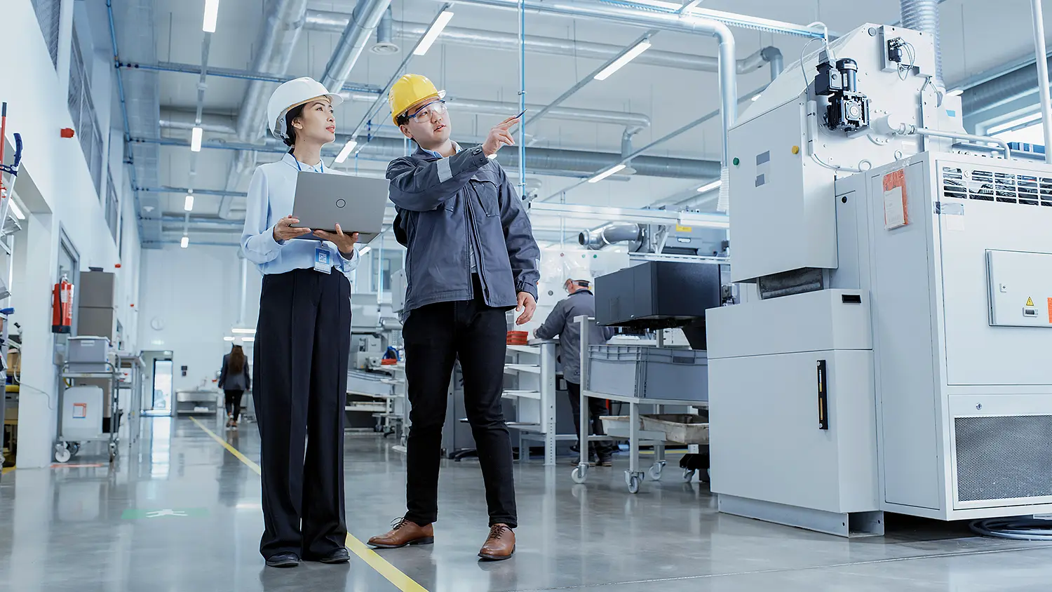 Two people are standing in a factory hall. One person is explaining something to the other and pointing to the left. 