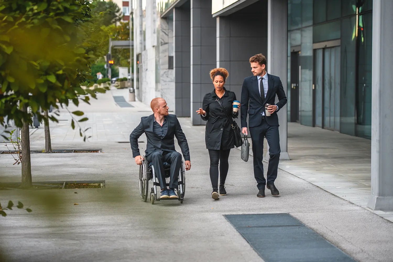 Three people – one in a wheelchair – are walking and talking on the sidewalk in front of a new building.