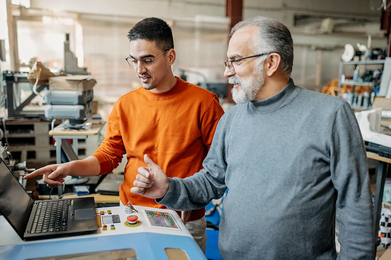 In a factory hall, a younger man and an older man are standing at a control unit with a laptop.