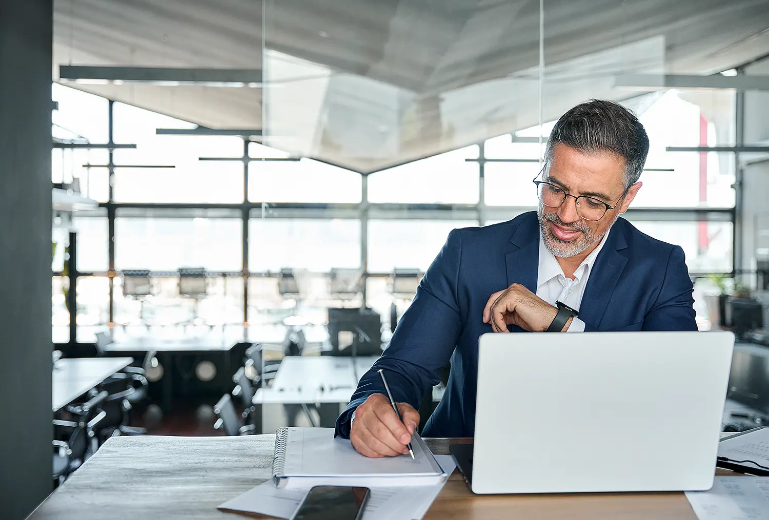 Ein Mann 50+ sitzt an einem Laptop und schreibt mit rechts etwas auf einen Schreibblock.